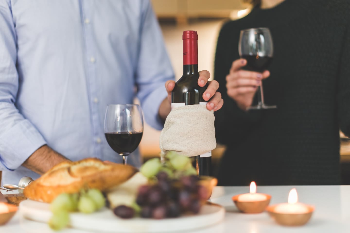 Un tavolo apparecchiato con pane, uva, formaggio e candele accese; dietro, due persone versano e tengono in mano calici di vino rosso.