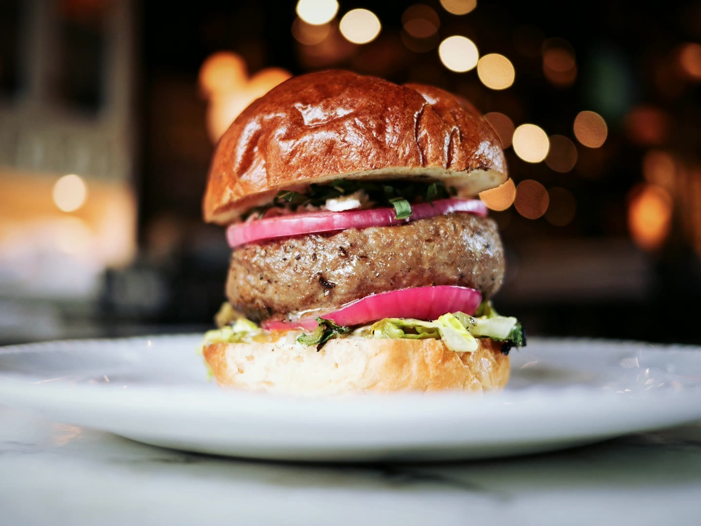 Un hamburger con cipolla rossa e insalata in un panino lucido, su un piatto bianco, davanti a luci calde sfocate.