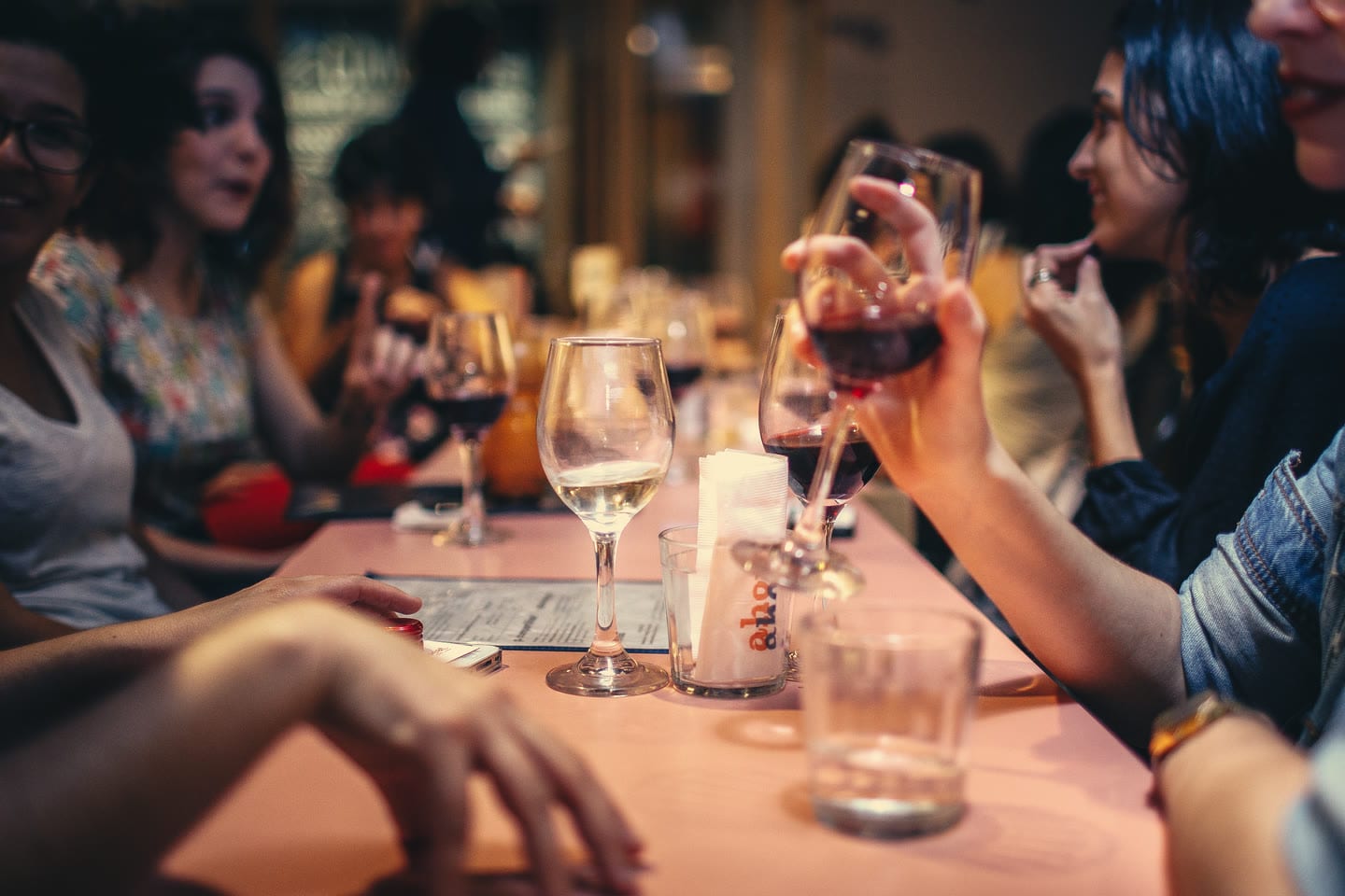 Un gruppo di amici a un lungo tavolo di un locale, la sera, che chiacchierano con i calici di vino in mano.