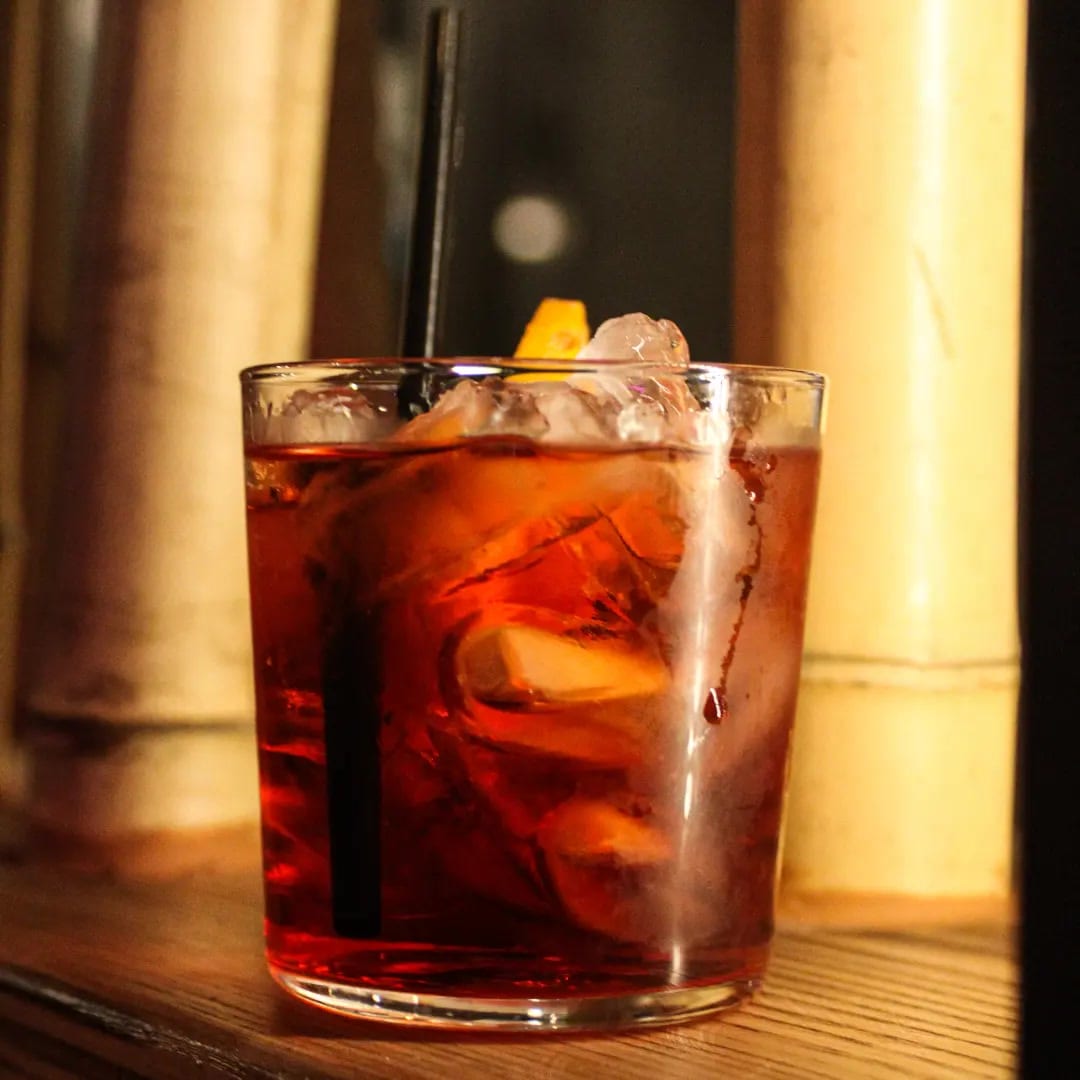 Un Negroni in un tumbler basso con ghiaccio e scorza d'arancia, su un ripiano in legno con luce calda.