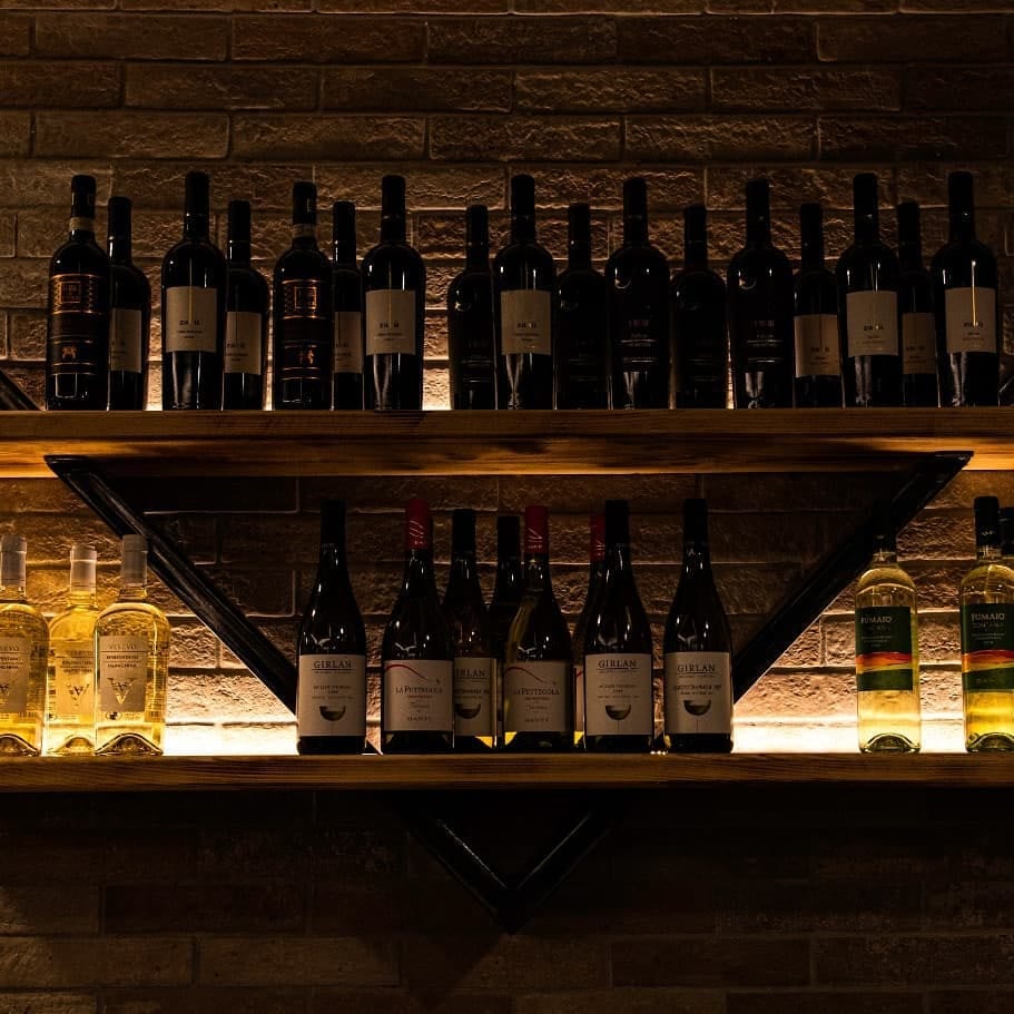 Mensole in legno e ferro cariche di bottiglie di vino rosso e bianco, illuminate dal basso contro un muro in mattoncino.