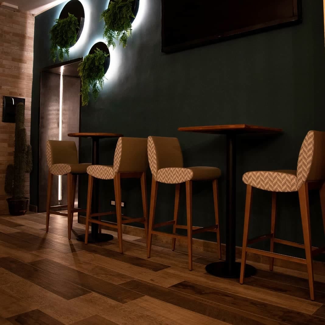Sgabelli alti in legno e tessuto chevron attorno a tavolini, sotto la parete scura con i dischi luminosi e le piante pendenti.